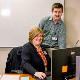 Two Monadnock Community Hospital employees, a woman seated at a computer and a man standing behind her, smile while working together in an office with a whiteboard in the background.