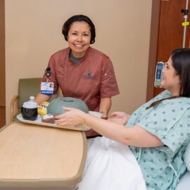 A food services team member at Monadnock Community Hospital delivers a meal tray to a patient sitting up in bed, both smiling warmly.