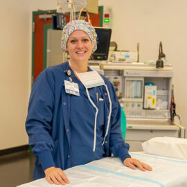 A surgical nurse in scrubs and a colorful cap smiles while standing at a prep table in an operating room at Monadnock Community Hospital.