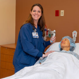 A nurse checks on a patient in pre-op at Monadnock Community Hospital, holding a thermometer and smiling as the patient looks up from the hospital bed.