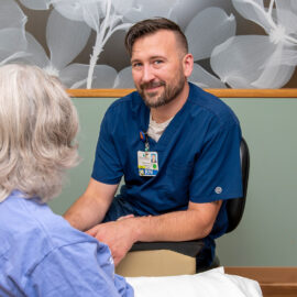 A male registered nurse kneels beside a seated patient, offering reassuring care in a calm, private setting at Monadnock Community Hospital.