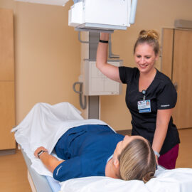 A radiologic technologist assists a patient during an imaging procedure at Monadnock Community Hospital, adjusting equipment while smiling.