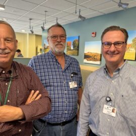 Three Monadnock Community Hospital engineering team members smile together in a common area, with framed landscape photos on the wall behind them. All are wearing collared shirts and employee badges, celebrating Hospital Week.