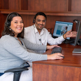 Two IT team members at Monadnock Community Hospital wear headsets and work together at a dual-computer setup, smiling toward the camera.