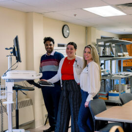 Three Monadnock Community Hospital staff members stand at a mobile computer cart in a shared workspace, smiling in a collaborative environment.