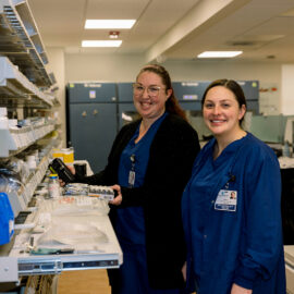 Two pharmacy techs work at a medication dispensing area with shelves.