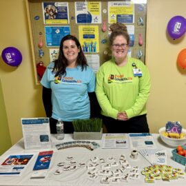 Two Monadnock Community Hospital staff members smile behind a colorful Nutrition Services table display for National Nutrition Month. The table features educational materials, healthy snacks, buttons, and giveaway items. A bulletin board behind them highlights colorectal cancer screening awareness.