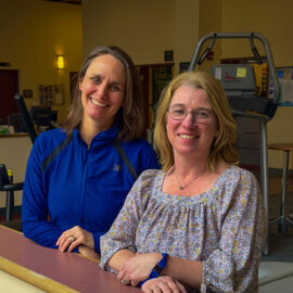 Two Monadnock Community Hospital occupational therapists pose together at the rehab gym. One wears a blue athletic jacket and the other a floral blouse, both smiling in front of therapy equipment.