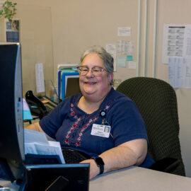 A smiling employee sits behind a computer at a desk surrounded by files and forms.