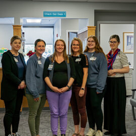 Six team members from Monadnock Orthopaedic Associates stand together in a clinic hallway under a sign labeled “Check Out Desk 1.” The group smiles warmly, wearing professional attire and ID badges.