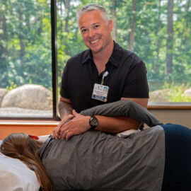 A physical therapist at MCH smiles while assisting a patient with shoulder mobility. The session takes place in a bright room with large windows overlooking a forest.
