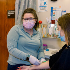 A masked phlebotomist at Monadnock Community Hospital draws blood from a patient in a clinical exam room. She wears gloves, a light blue top, and pink mask, and smiles while providing care.
