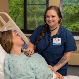 A nurse at Monadnock Community Hospital’s Birthing Suite listens to a patient's heartbeat with a stethoscope. The nurse wears navy scrubs and a braid, smiling at the camera while the patient lies in bed.