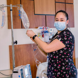 A nurse in black scrubs adjusts an IV line for a patient in a hospital room at MCH. She wears a mask and patterned scrub top and is surrounded by clinical equipment.
