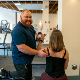 A physical therapist at Monadnock Community Hospital kneels beside a patient, guiding her through a shoulder mobility exercise in the rehab gym. The therapist smiles while engaging the patient.