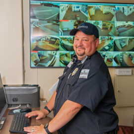 A Monadnock Community Hospital security officer stands at a computer station in the security office, smiling at the camera. A wall of surveillance monitors shows views from around the hospital.