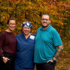 Three Monadnock Community Hospital employees smile outdoors against a vibrant backdrop of autumn foliage. One wears navy blue scrubs and a floral surgical cap, standing between two casually dressed colleagues.