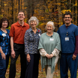 Five Monadnock Community Hospital team members stand together in front of a forest filled with fall colors. They are smiling and wearing a mix of professional and clinical attire.