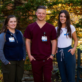 Three young Monadnock Community Hospital staff members stand outside in dappled sunlight. One wears burgundy scrubs, while the others wear casual and professional clothing with visible ID badges.