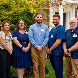 Five MCH employees stand smiling in front of the hospital entrance, framed by lush greenery. The group includes both clinical and non-clinical team members dressed in professional attire.