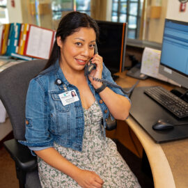 A switchboard operator smiles while speaking on the phone at her desk at Monadnock Community Hospital. She wears a floral dress and denim jacket, with two monitors in front of her.