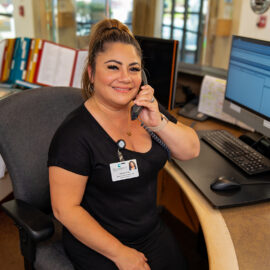 A switchboard operator at MCH smiles warmly while on the phone. She wears a black dress and sits at a workstation with dual monitors and hospital communication tools.
