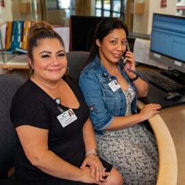 Two switchboard operators at Monadnock Community Hospital sit side-by-side at their desk. One is on the phone while the other smiles at the camera, both surrounded by computer monitors and paperwork.