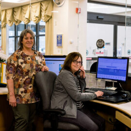 Two MCH team members work behind a glass window at the hospital switchboard. One is standing and smiling, while the other is seated and speaking on the phone.