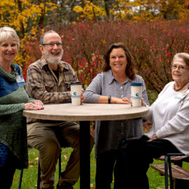 our Monadnock Community Hospital volunteers enjoy coffee together at an outdoor round table surrounded by colorful fall foliage. They are smiling and wearing volunteer or staff badges.