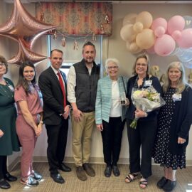 Laura Moran, RN, stands holding a bouquet and award surrounded by smiling Monadnock Community Hospital colleagues and leaders at a celebration to receive the 2025 Clint Jones Nursing Award. Pink and gold balloons decorate the background.