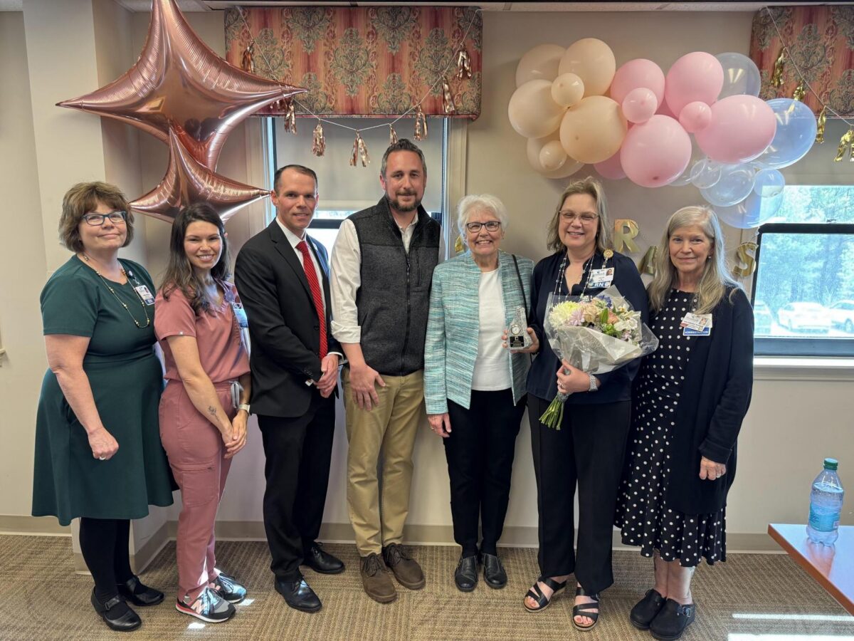 Laura Moran, RN, stands holding a bouquet and award surrounded by smiling Monadnock Community Hospital colleagues and leaders at a celebration to receive the 2025 Clint Jones Nursing Award. Pink and gold balloons decorate the background.