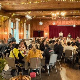 Emergency medical personnel and hospital staff gather at decorated round tables inside a warmly lit, rustic wood-paneled hall for Monadnock Community Hospital’s 2025 EMS Recognition Dinner, with string lights and snowflake decorations overhead.