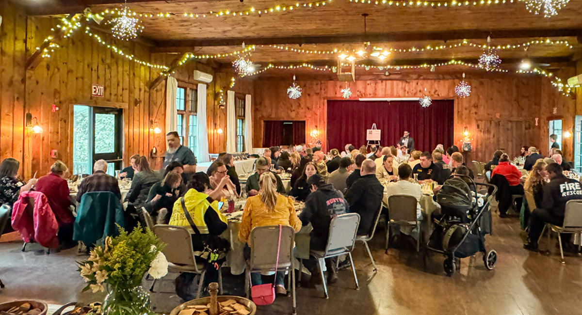 Emergency medical personnel and hospital staff gather at decorated round tables inside a warmly lit, rustic wood-paneled hall for Monadnock Community Hospital’s 2025 EMS Recognition Dinner, with string lights and snowflake decorations overhead.