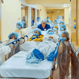 A group of children dressed in surgical gowns, masks, and hair covers push a hospital bed down the hallway at Monadnock Community Hospital during a Girl Scouts visit. A staff member in scrubs smiles behind them, guiding the playful, educational activity.