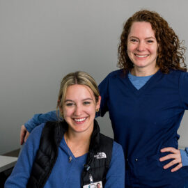Two Monadnock Community Hospital employees, one standing and one seated, smile together in a casual office setting with gray walls and simple furnishings.