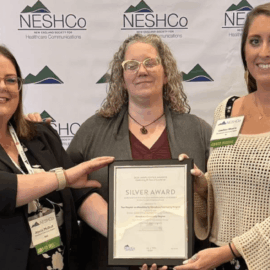 Three Monadnock Community Hospital staff members smile and proudly hold a framed Silver Award certificate in front of a NESHCo (New England Society for Healthcare Communications) backdrop, celebrating their Lamplighter Award win for the “Your Hospital” eNewsletter.