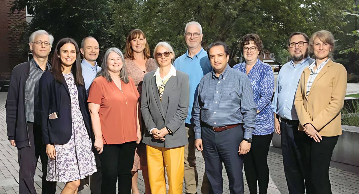 A group of 11 professionally dressed adults standing outdoors on a paved walkway, posing for a photo with trees and buildings in the background. The group includes Dr. Shawn Harrington, seen in a blue shirt and khaki pants near the rear center.