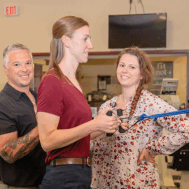 Two physical therapists assist a patient using a resistance band in a spacious rehab gym, all smiling and engaged.