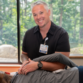 A physical therapist at MCH smiles while assisting a patient with shoulder mobility. The session takes place in a bright room with large windows overlooking a forest.