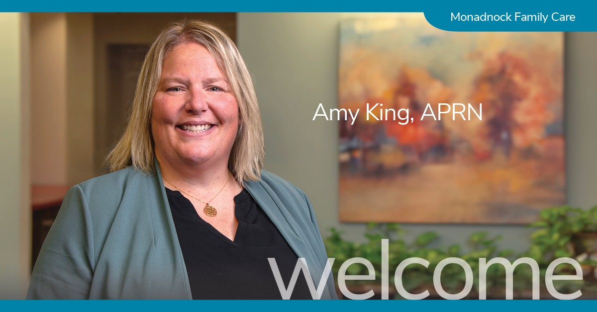 Smiling woman with blonde hair wearing a sage green jacket over a black top, standing indoors with an abstract autumnal painting in the background. Text reads “Amy King, APRN” with a banner that says Monadnock Family Care.