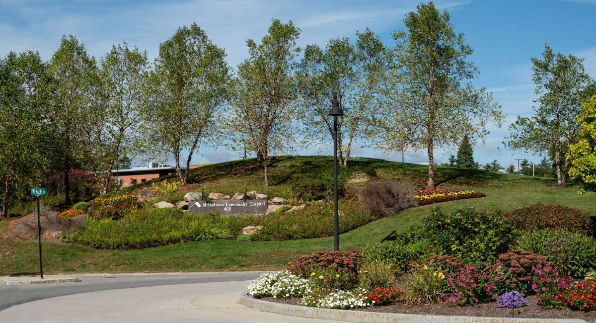 Entrance to Monadnock Community Hospital landscaped with flowers and a large red banner announcing the 2025 Keene Sentinel Choice Awards with a silver star emblem.