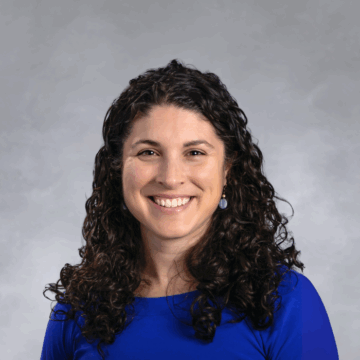 Smiling woman with long, dark curly hair wearing a royal blue top and dangling earrings, in front of a soft gray studio background.