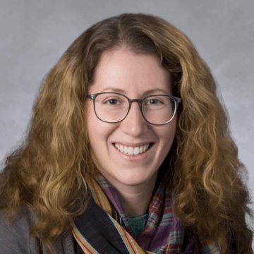 Smiling woman with long, curly light brown hair and glasses, wearing a multicolored scarf and gray top, in front of a soft gray studio background.
