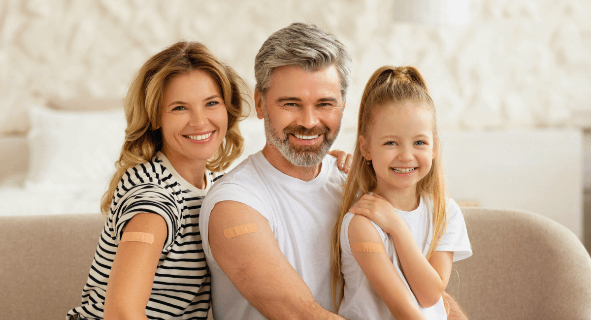 A family of three—mother, father, and young daughter—smile together on a couch with small adhesive bandages on their upper arms, indicating recent vaccinations. On the right, a blue panel features a circular headshot of Brian Earnshaw, RN, BSN, with the text: "A note from our Infection Preventionist, Brian Earnshaw, RN, BSN." The image emphasizes family health and professional vaccine guidance.