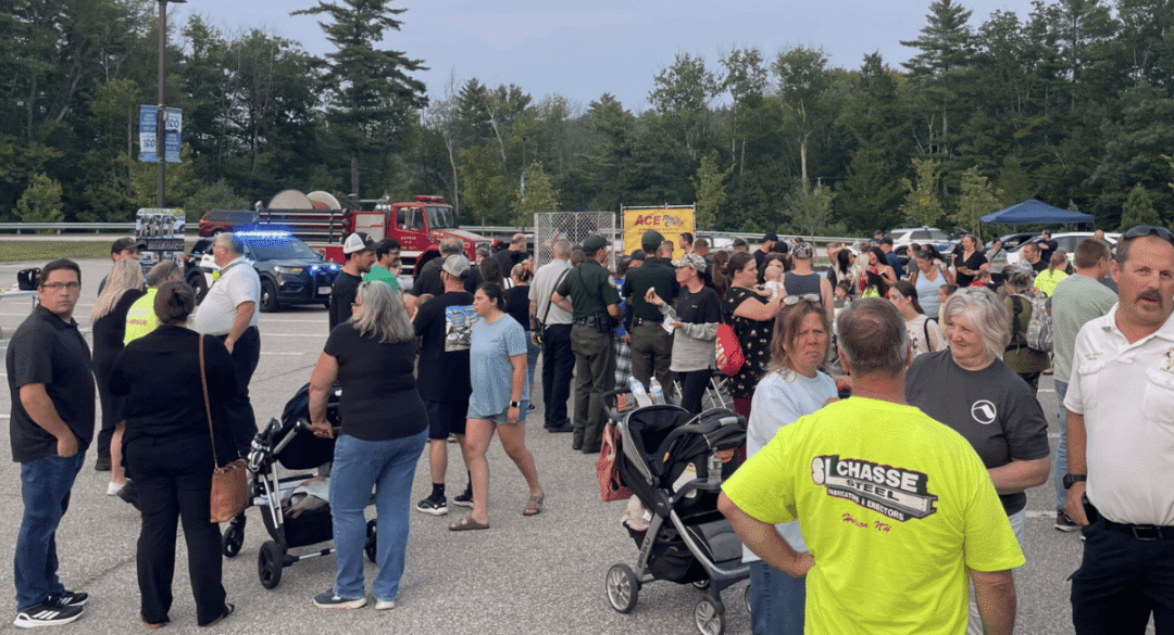 arge crowd gathered at an outdoor community event in a parking lot, with people talking, pushing strollers, and mingling. A police car with flashing lights and a fire truck are visible in the background, along with event booths and tents set up under a cloudy evening sky.