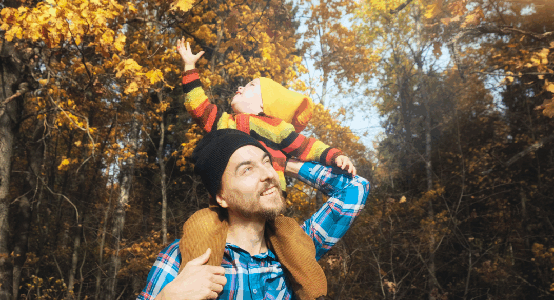 A smiling man in a plaid shirt carries a young child on his shoulders during an autumn walk. The child, dressed in a colorful striped sweater and orange hat, reaches toward golden leaves on a tree overhead.