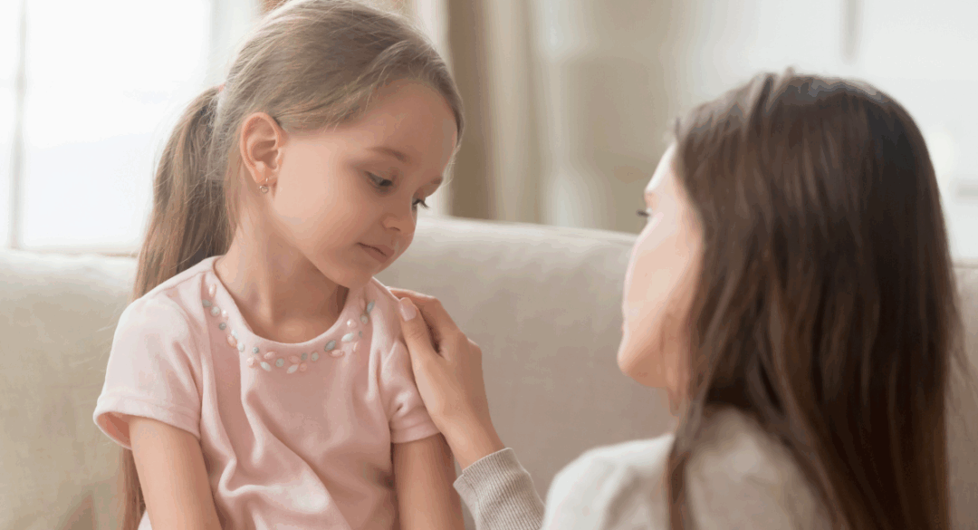 A young girl in a pink shirt looks down sadly while sitting on a couch as another person gently places a hand on her shoulder in reassurance.