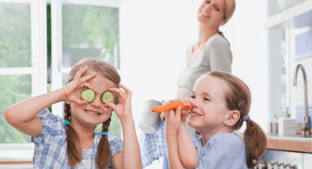 Two young girls playfully hold vegetables while helping in the kitchen, one holding cucumber slices over her eyes and the other pretending a carrot is a trumpet, with fresh produce on the counter and an adult smiling in the background.