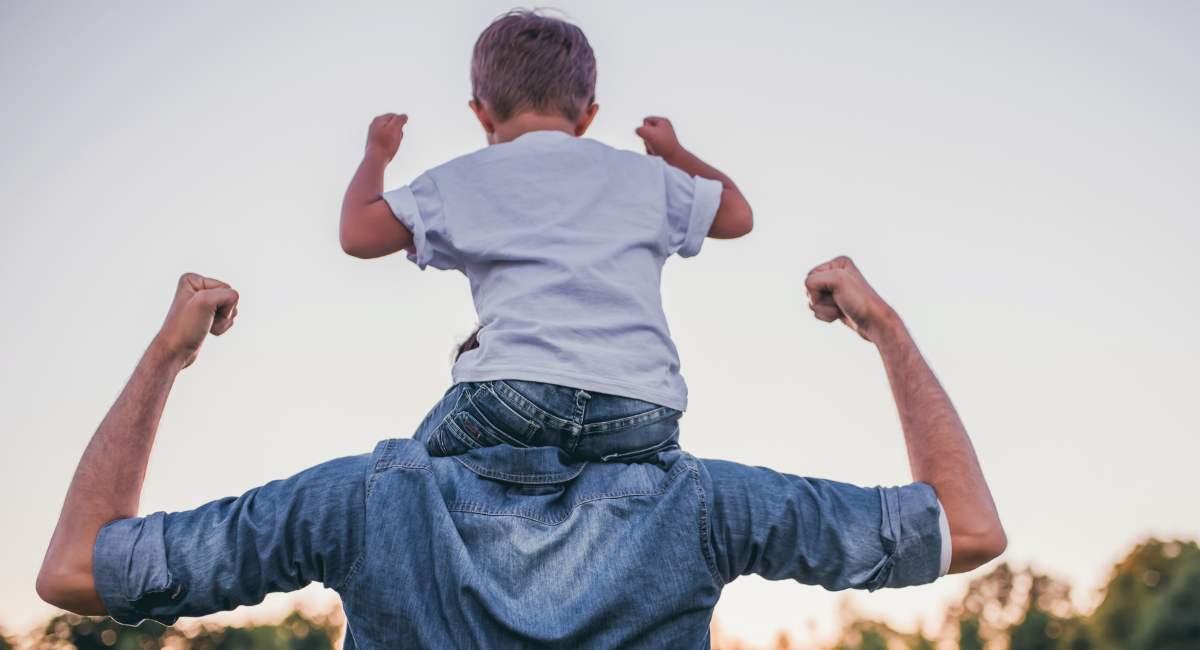 A boy sits on an adult’s shoulders outdoors at sunset, both raising their arms in a flexing pose, symbolizing strength and support.