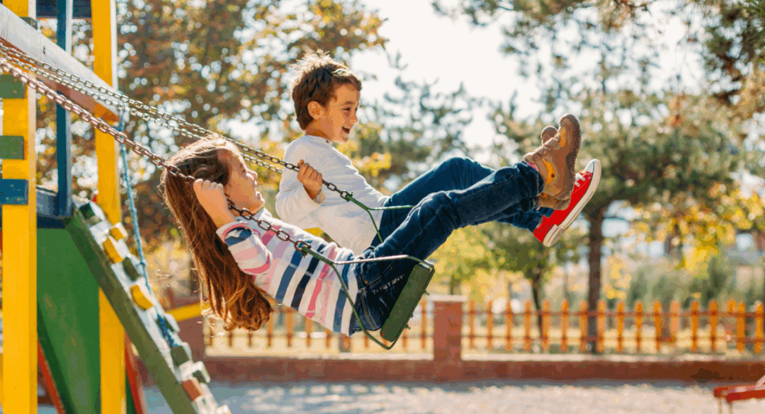 A boy and girl laugh together while swinging side by side on a playground swing set on a sunny day.